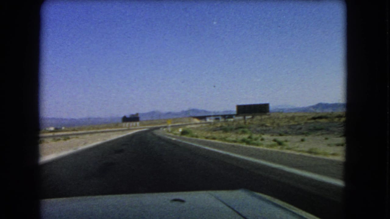 1966 Nevada highway perspective from a moving vehicle, with open desert, roadside signs, and mountains ahead.