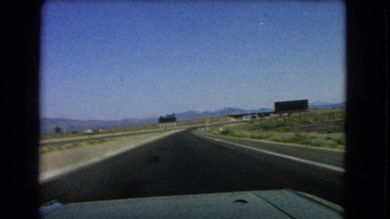 1966 8mm view from a moving car on a curving desert road near Las Vegas, with distant mountains and signs ahead.
