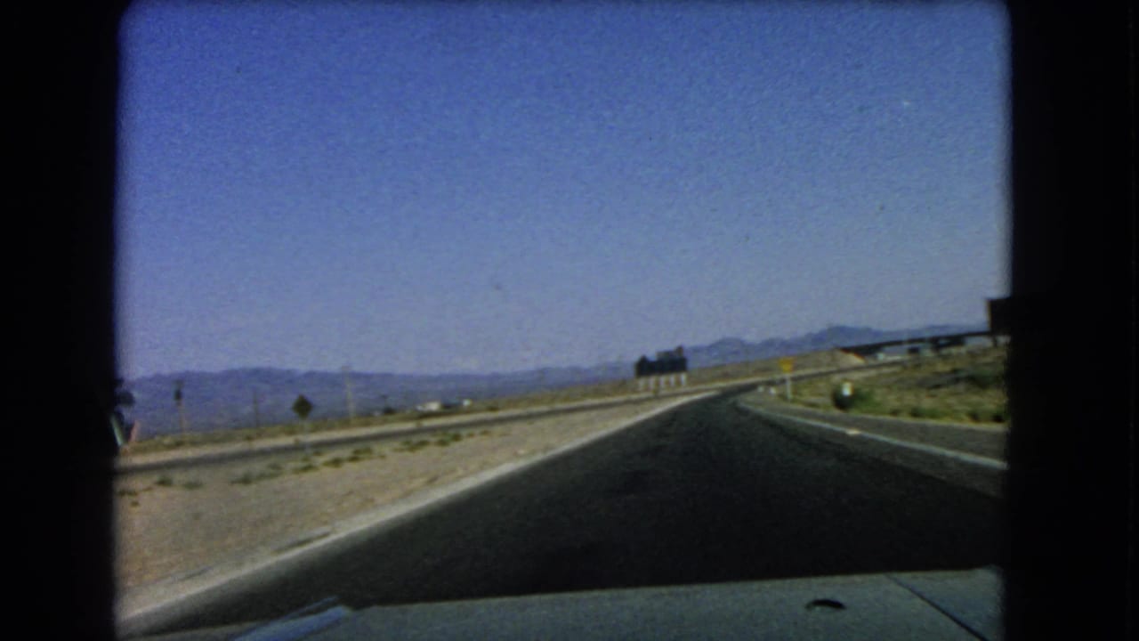 Roadway curves right through desert terrain with low mountains and signs in the distance.
