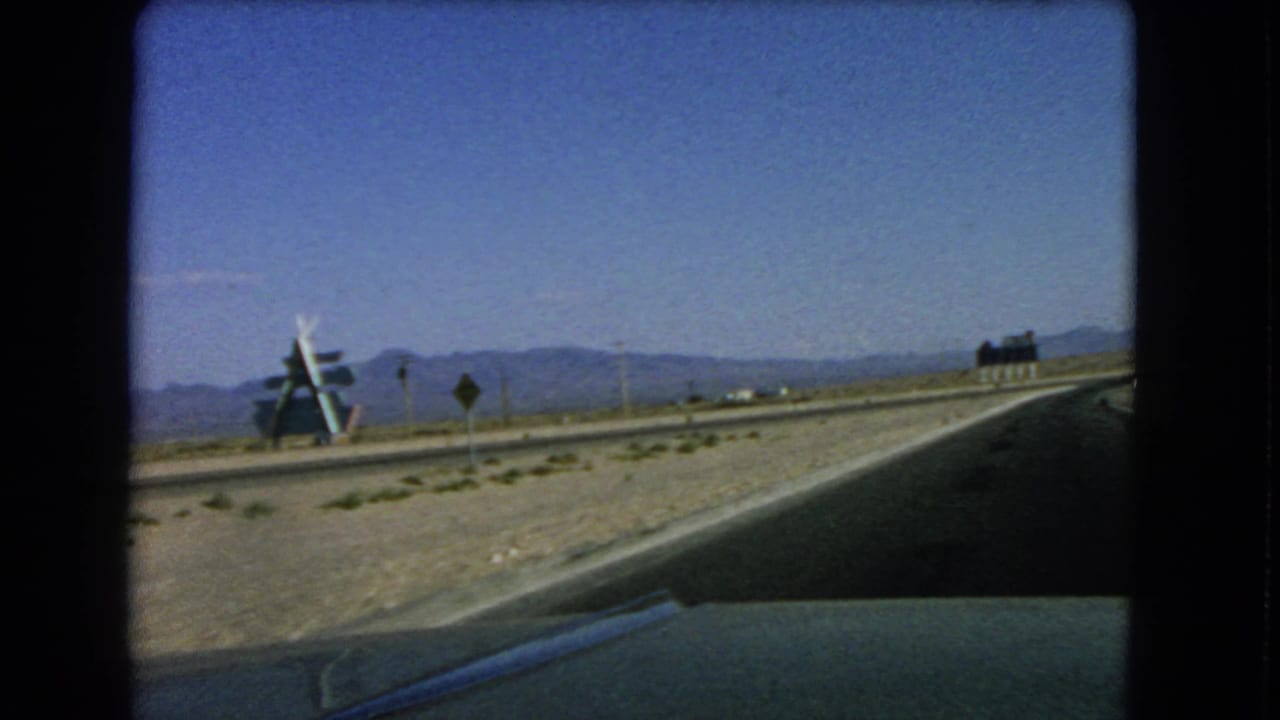 1966 home-movie footage shows a moving-car view of a Nevada desert road with signs, mountains, and an angled roadside structure.