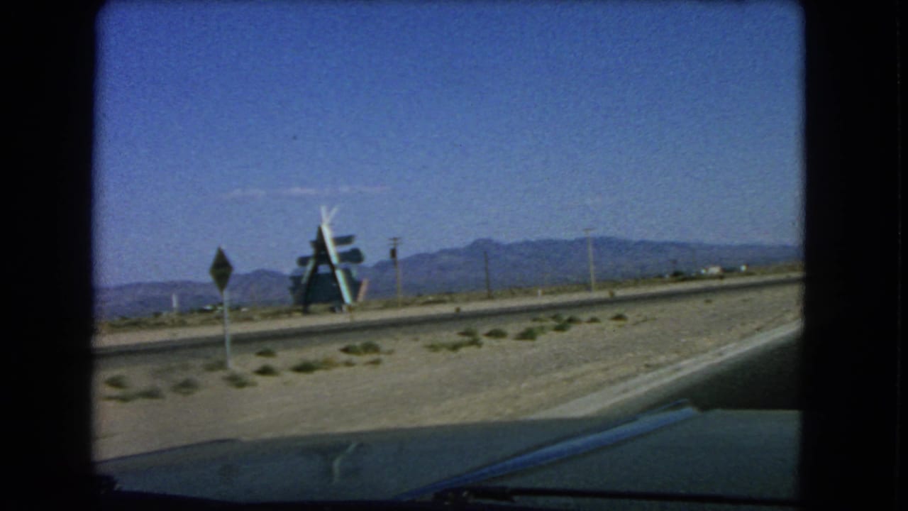 1966 home-movie frame from a moving vehicle showing a road sign, roadside attraction-like structure, low desert plants, and mountains near Las Vegas.