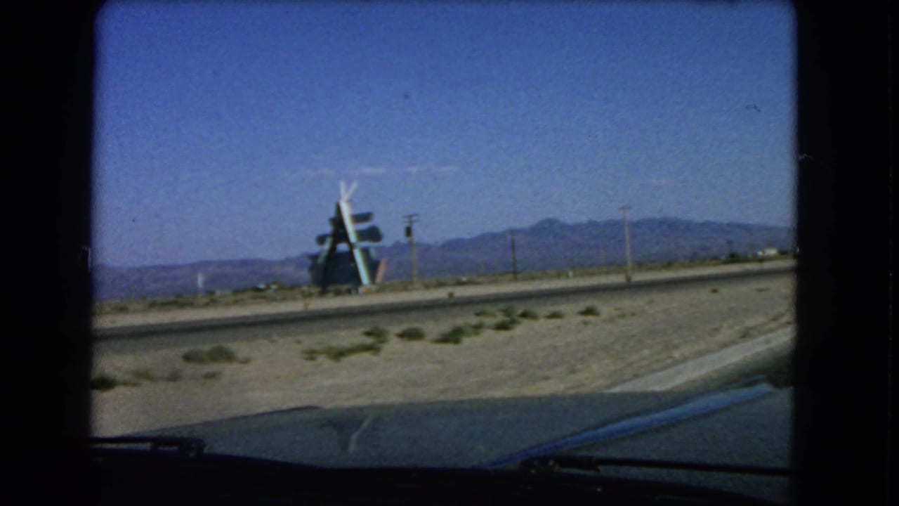 Las Vegas-area 1966 8mm view of a roadside structure across a desert road, framed by a car windshield, utility poles, and a distant mountain range.