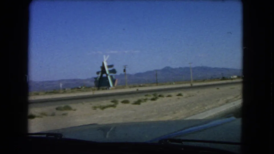 Las Vegas-area 1966 8mm view of a roadside structure across a desert road, framed by a car windshield, utility poles, and a distant mountain range.
