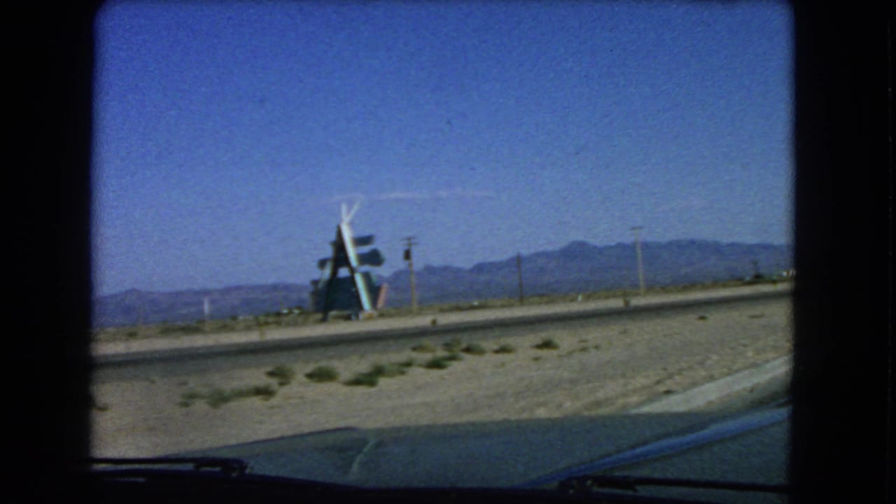 1966 Las Vegas-area car-window view showing sandy roadside, a sign-like structure, utility poles, and distant desert mountains.