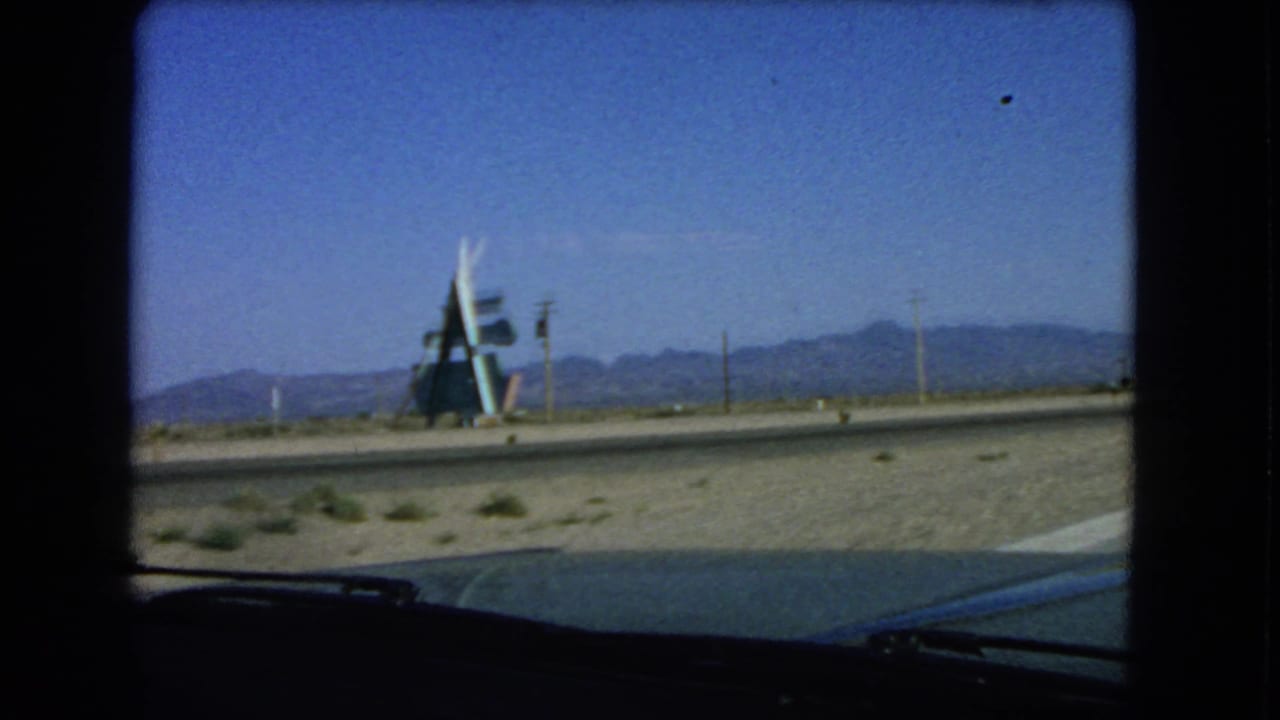 1966 Las Vegas-area windshield view of a desert roadside structure, open sand, low vegetation, and mountains under blue sky.