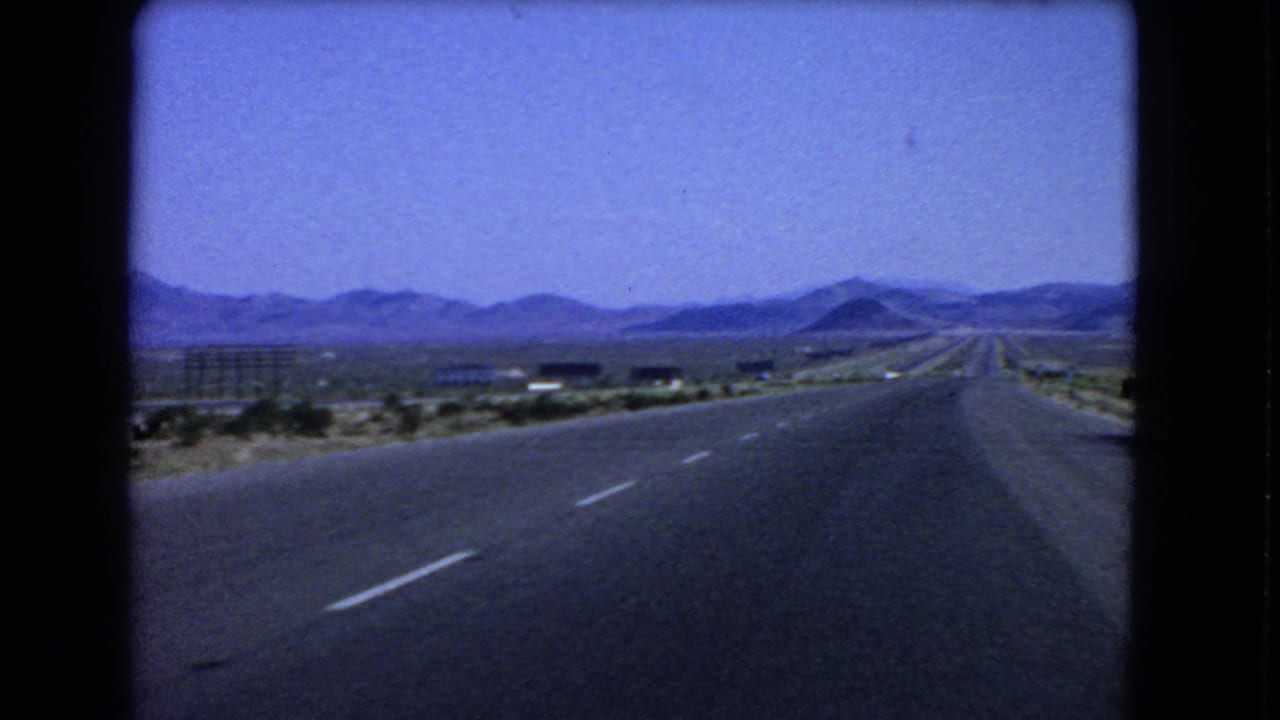 1966 Nevada desert highway view from inside a car, with the road leading toward low mountains and sparse roadside development.