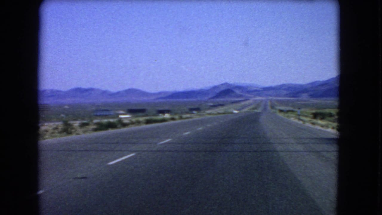 1966 Las Vegas-area road footage looking forward through the windshield at a straight highway, sandy shoulders, and distant mountains.