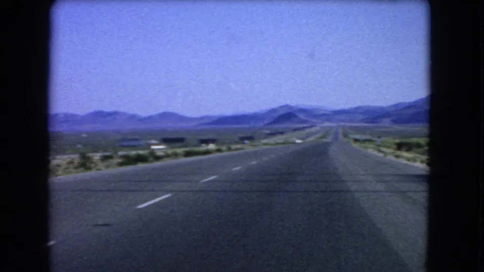 1966 Las Vegas-area road footage looking forward through the windshield at a straight highway, sandy shoulders, and distant mountains.