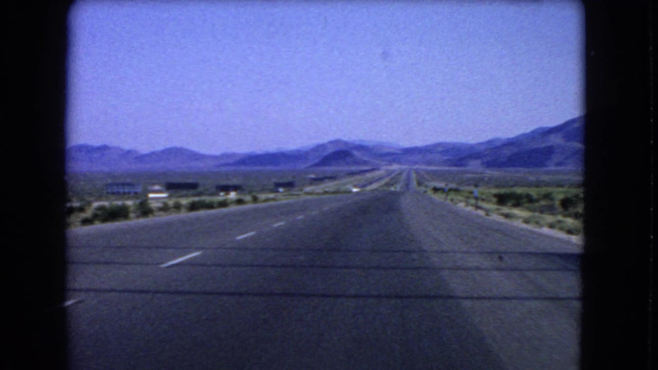 1966 8mm view near Las Vegas of a wide highway stretching through desert shrubs toward low mountains.