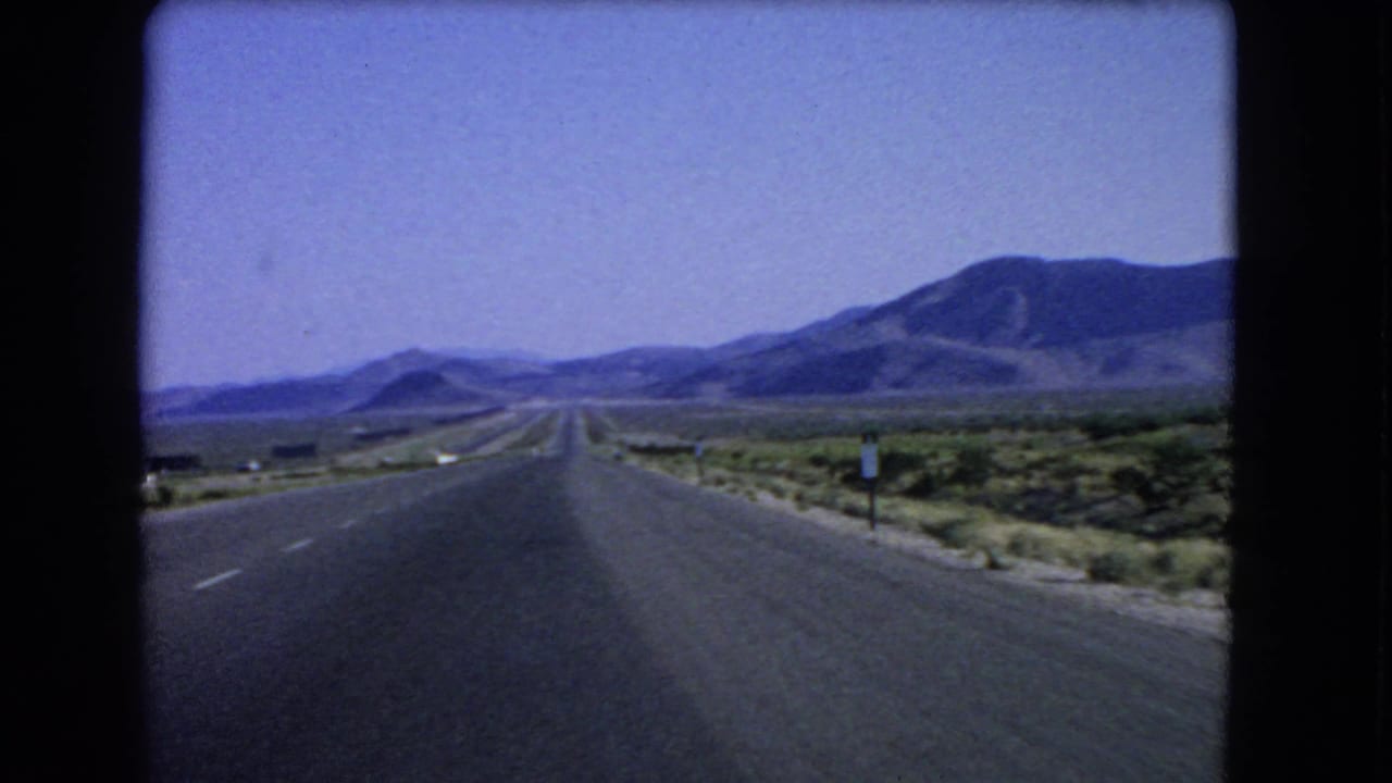 Nevada desert road scene from 1966 home-movie footage, with scrub vegetation and mountains ahead.