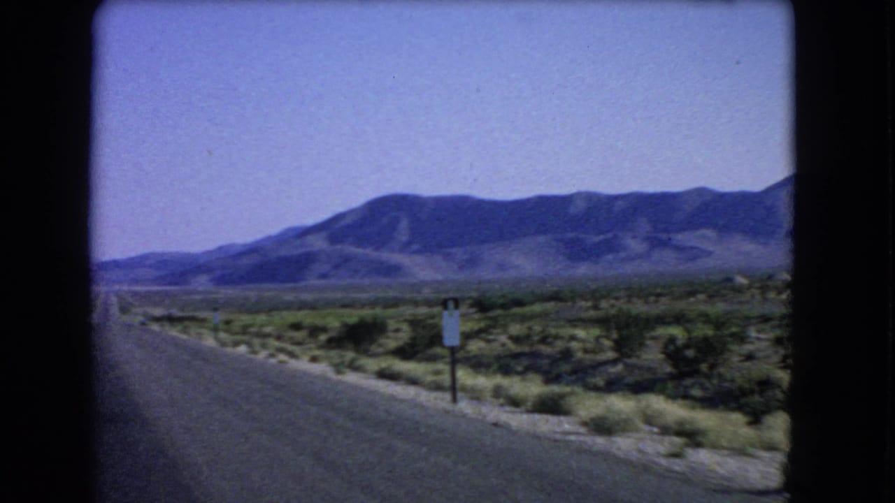1966 8mm frame near Las Vegas showing the highway edge, sparse desert plants, a roadside sign, and mountains.