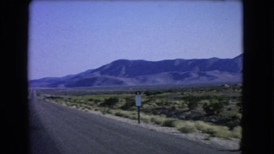 1966 8mm frame near Las Vegas showing the highway edge, sparse desert plants, a roadside sign, and mountains.