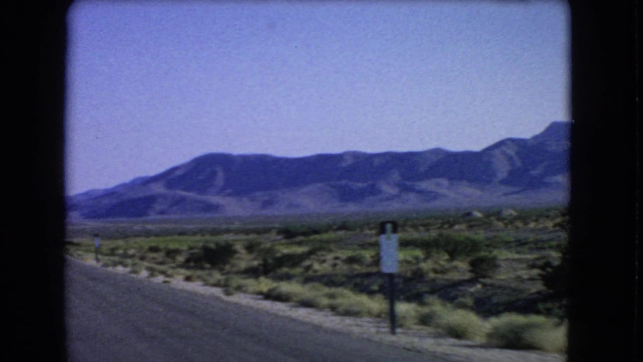 Roadside desert plain with shrubs, a small sign, and mountains under a blue sky.