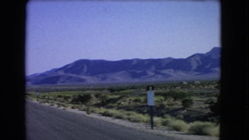 1966 8mm view near Las Vegas showing a paved road edge, desert shrubs, a roadside sign, and distant mountains.