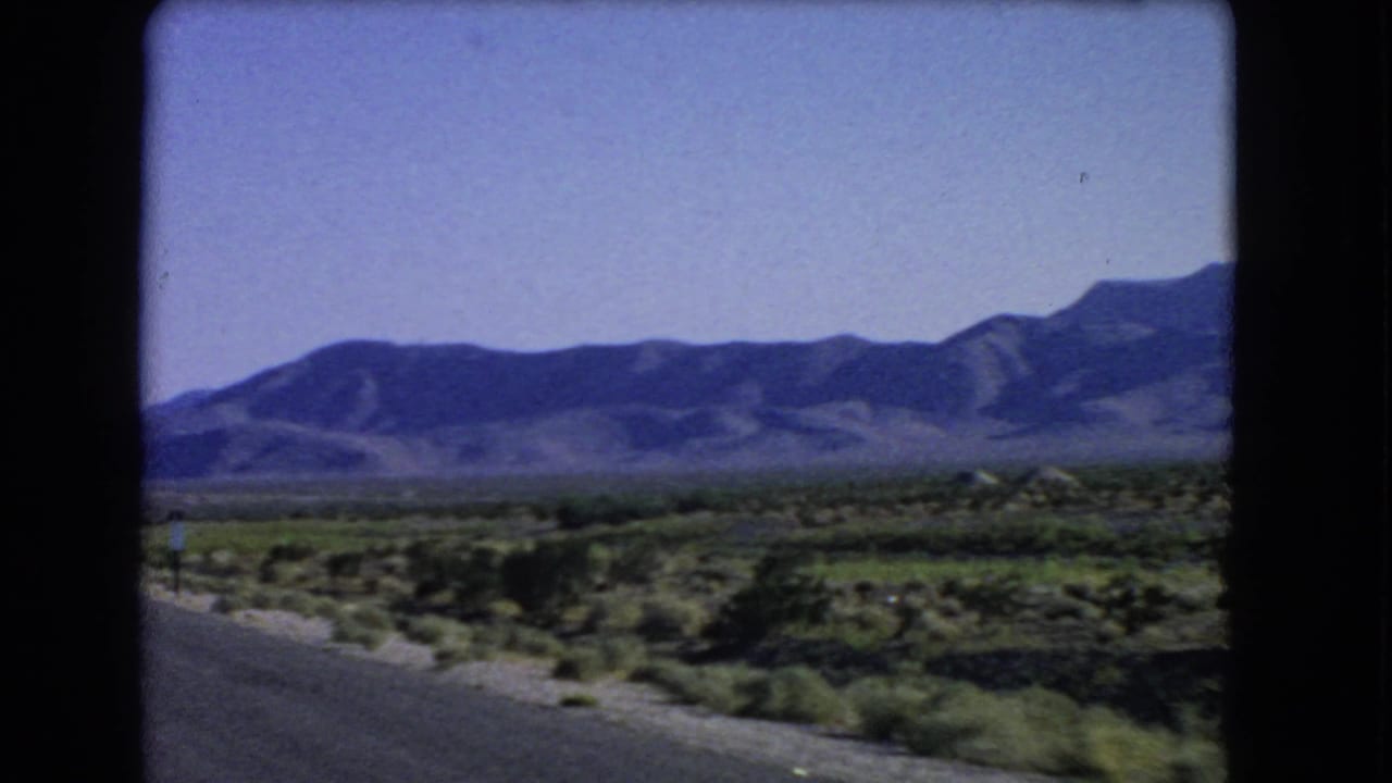 Desert road shoulder and scrubland lead toward a long mountain range.