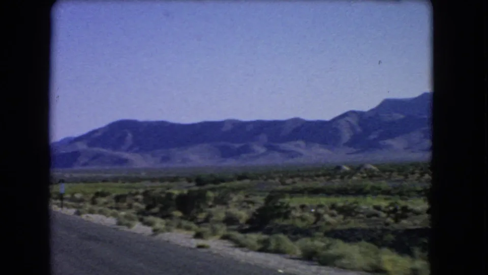 1966 8mm frame near Las Vegas with roadside desert vegetation, open terrain, and a blue mountain ridge beyond.