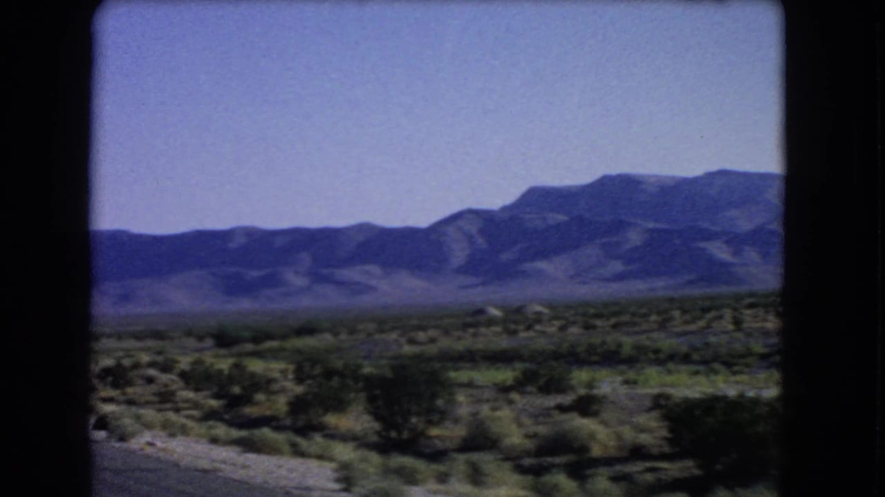 1966 8mm roadside view near Las Vegas, with motion-softened desert shrubs and mountains across the horizon.