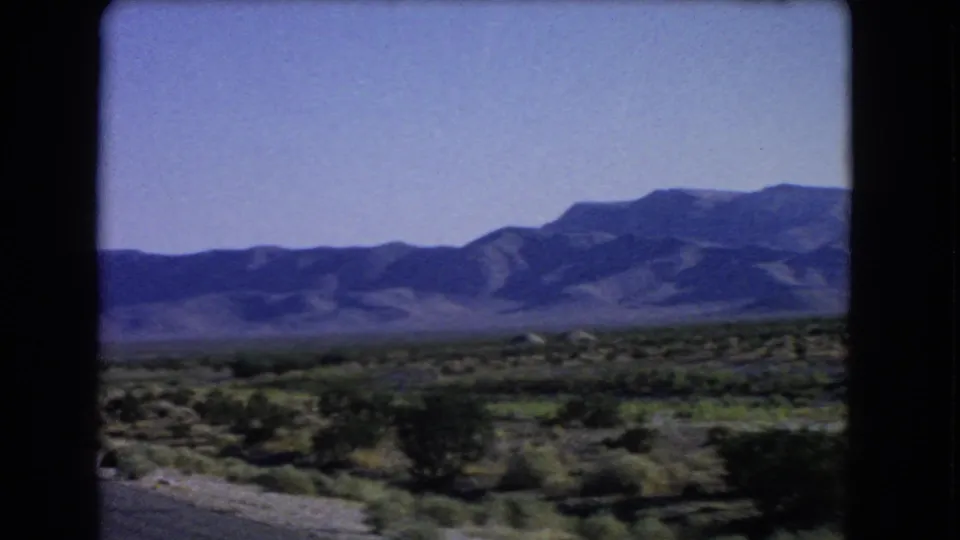 1966 8mm roadside view near Las Vegas, with motion-softened desert shrubs and mountains across the horizon.