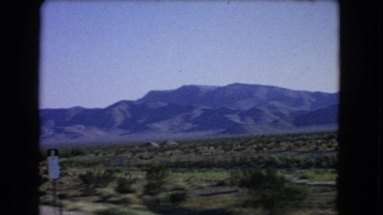1966 8mm view near Las Vegas showing a small roadside sign, desert scrub, open land, and mountains in the distance.