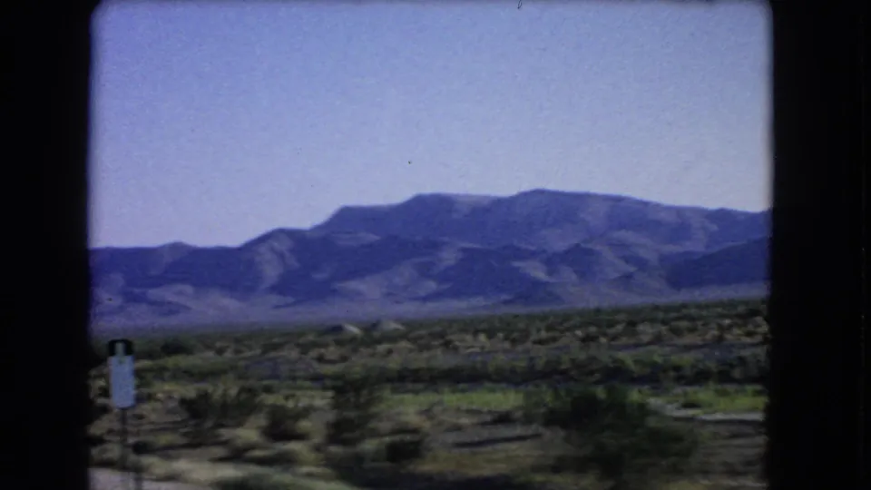 1966 8mm view near Las Vegas showing a small roadside sign, desert scrub, open land, and mountains in the distance.