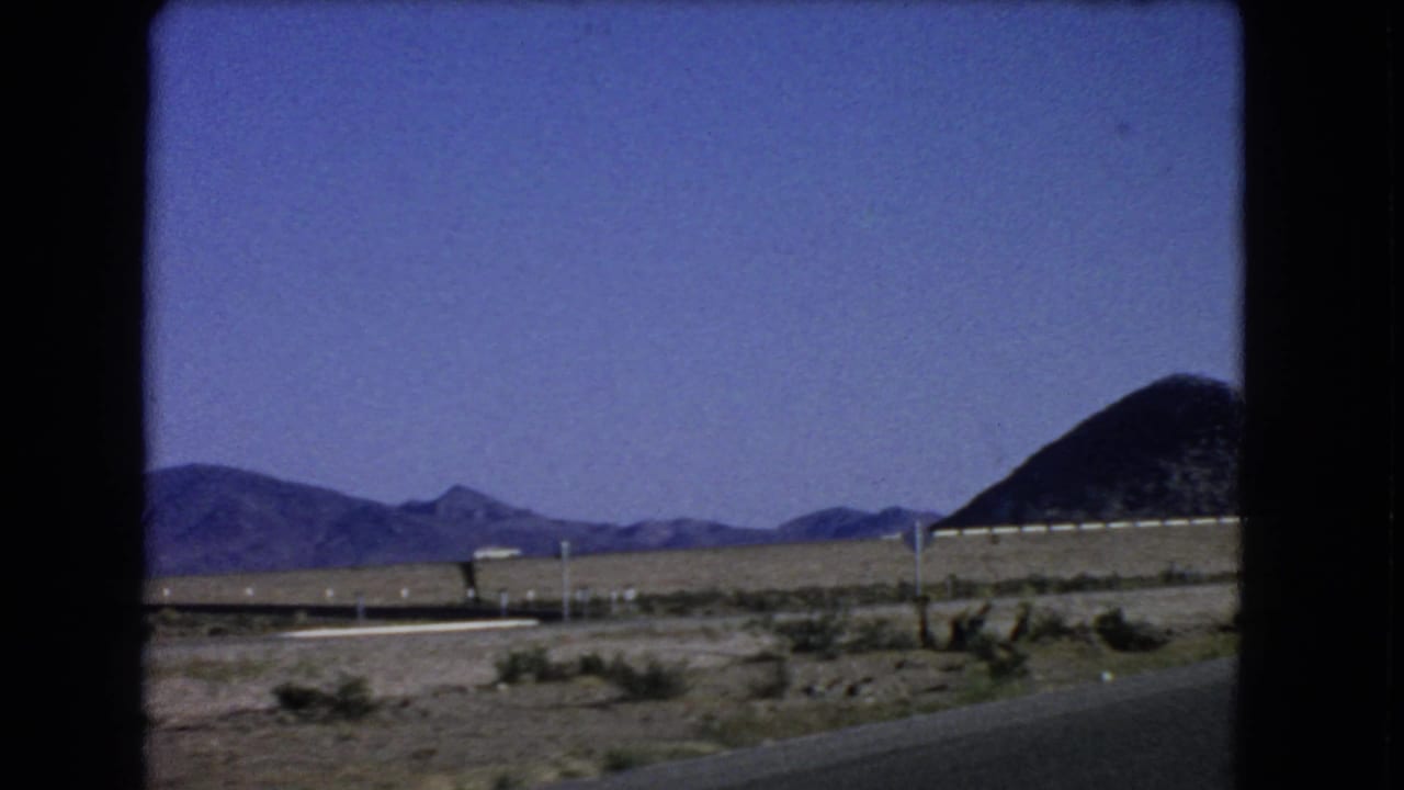 Car-window footage from 1966 near Las Vegas showing a paved road edge, desert scrub, roadside posts, and a dark hill.