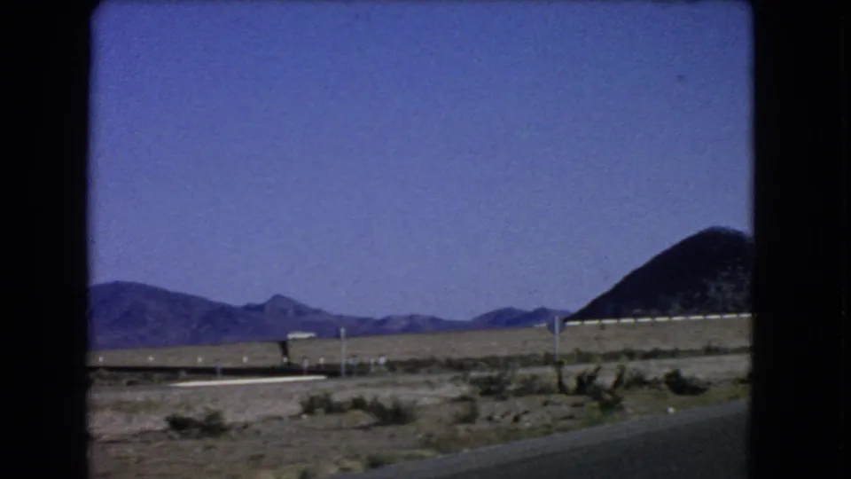 Car-window footage from 1966 near Las Vegas showing a paved road edge, desert scrub, roadside posts, and a dark hill.