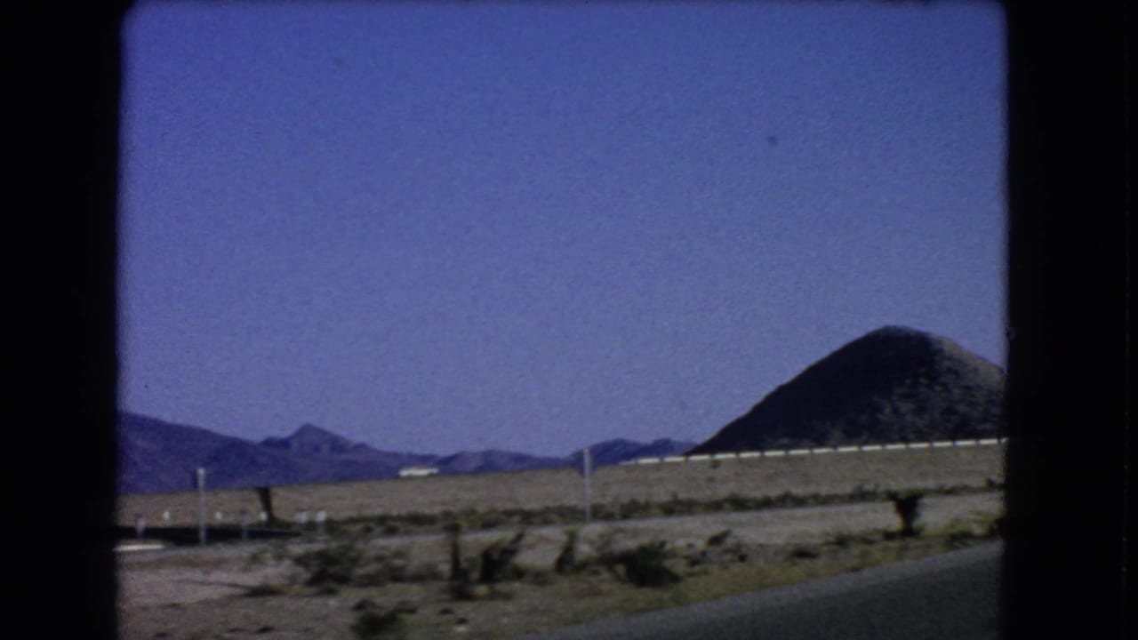 1966 8mm frame near Las Vegas with road shoulder, sparse brush, low mountains, and a dark rounded hill at right.