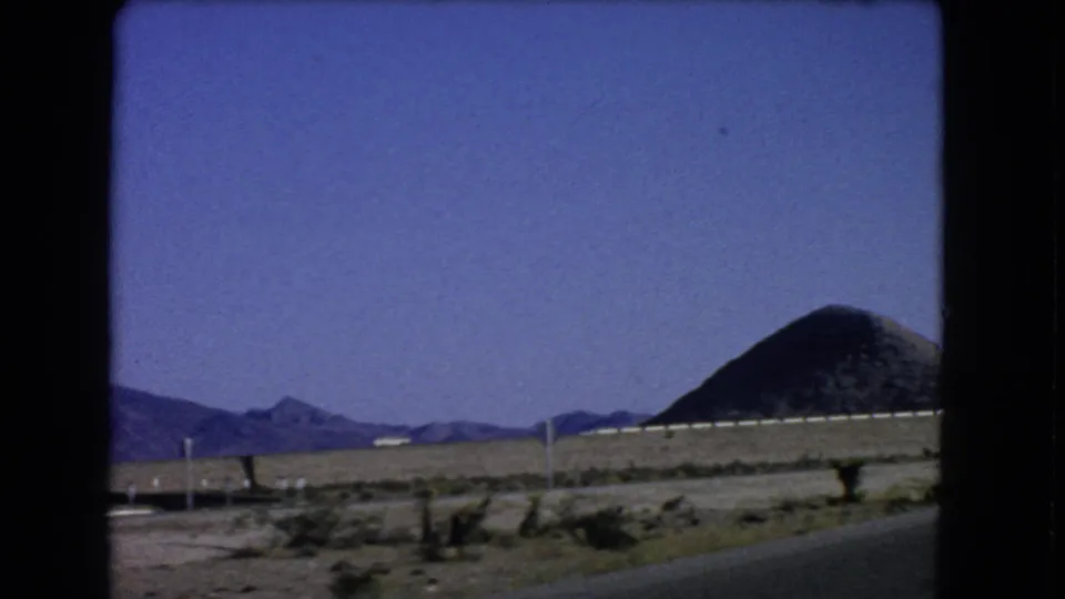 1966 8mm frame near Las Vegas with road shoulder, sparse brush, low mountains, and a dark rounded hill at right.