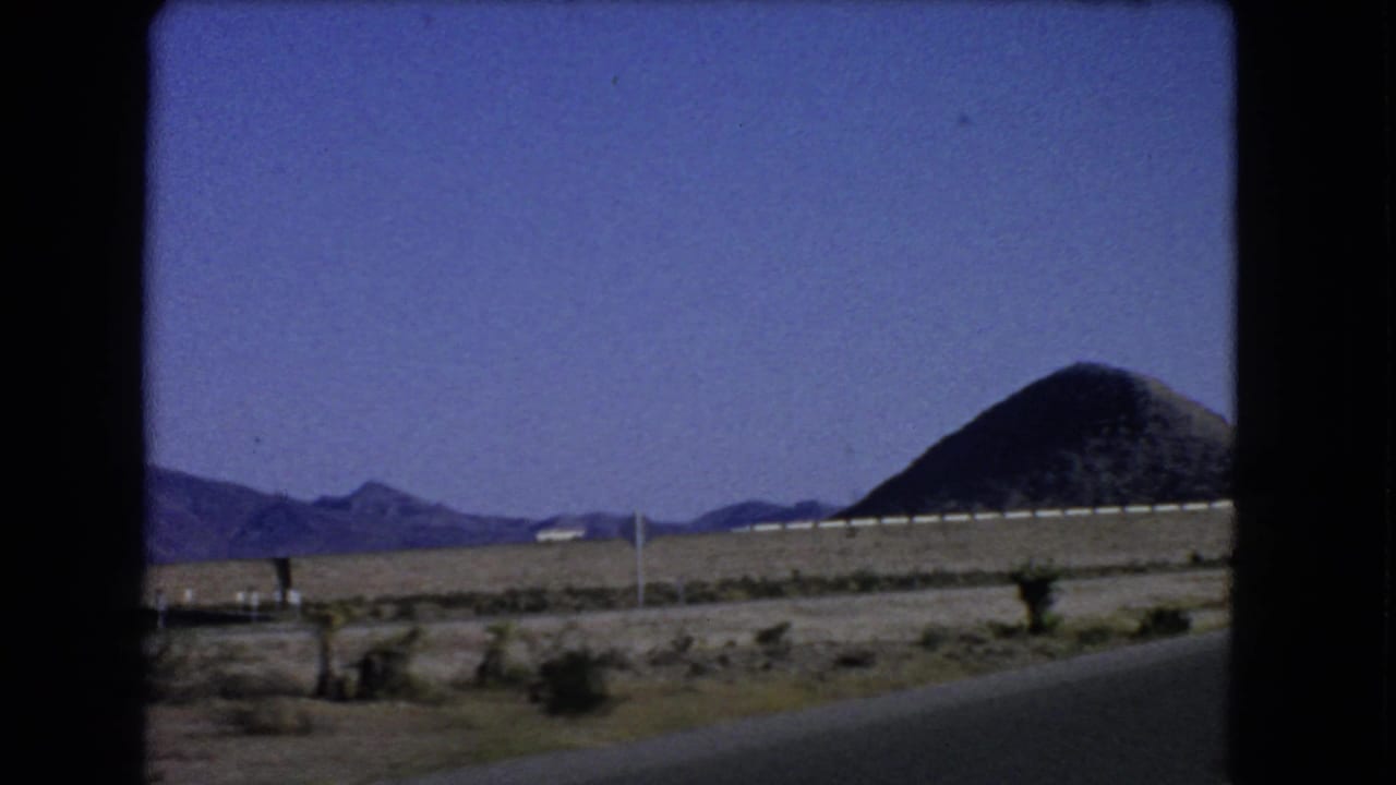 Desert road shoulder with distant mountains, utility poles, guardrail, and a conical hill.