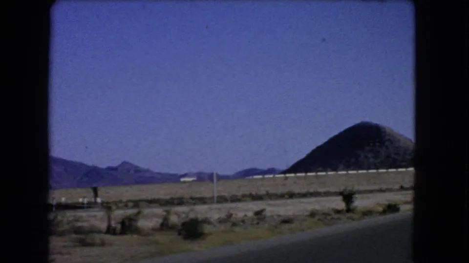 1966 8mm Nevada road-trip frame showing desert scrub, roadside poles, distant mountains, and a prominent conical hill near Las Vegas.