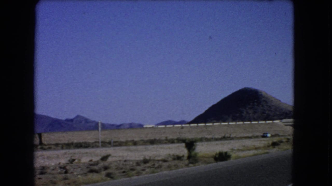 Roadside desert landscape with guardrail, mountains, utility poles, and conical hill.