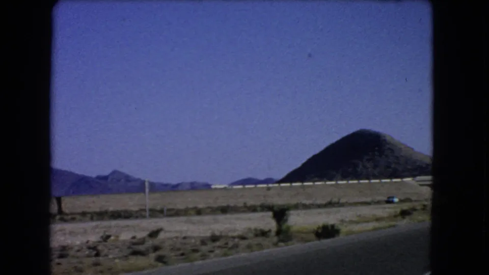 1966 Las Vegas, Nevada home-movie frame from a moving vehicle, with open desert, guardrail, distant ridges, and a dark conical hill.