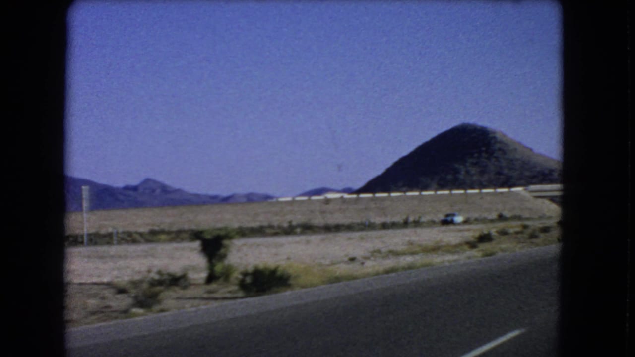 1966 8mm frame near Las Vegas showing a small blue car in the desert distance, with guardrail, road shoulder, mountains, and a conical hill.