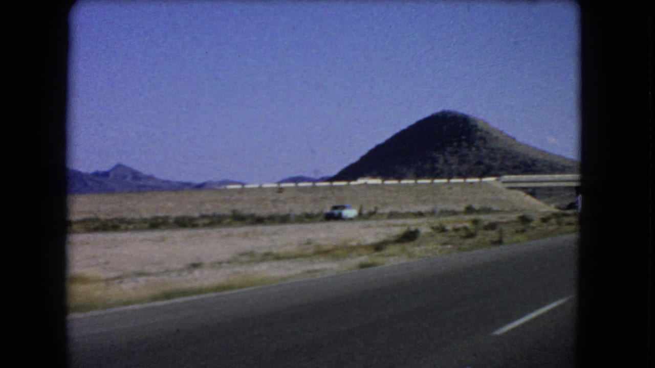 1966 Las Vegas, Nevada road-trip frame with asphalt in the foreground, a small blue car, desert scrub, a guardrail, mountains, and a conical hill.