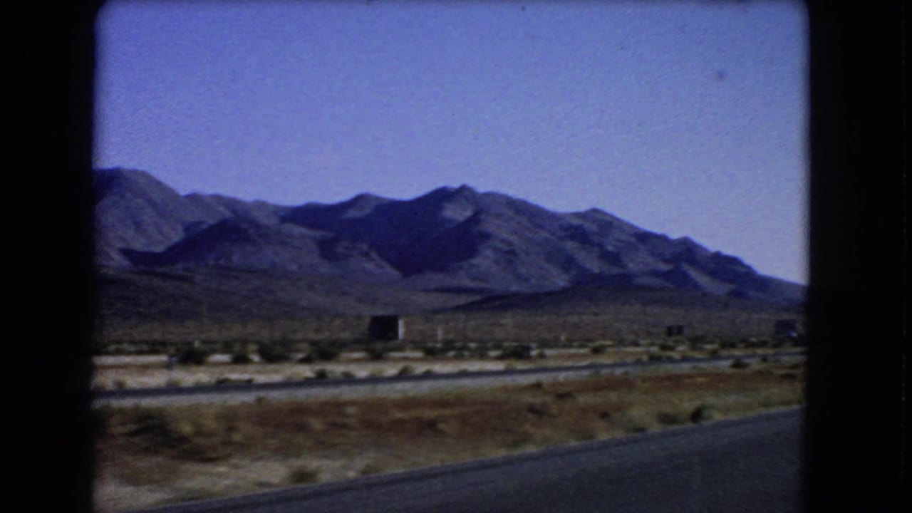 1966 Las Vegas-area 8mm frame showing a desert road edge, low scrub, and distant mountain ridges from a moving vehicle.