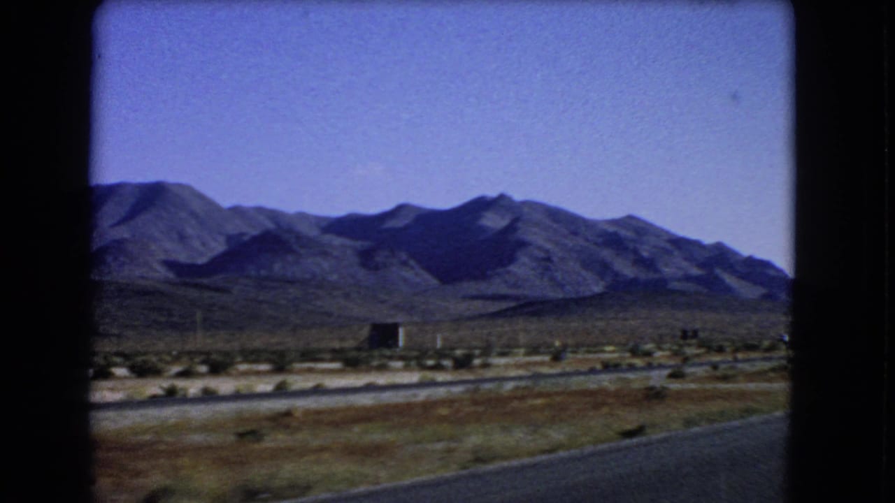 1966 Nevada home-movie frame with mountains, open desert ground, utility poles, and roadside pavement seen from a car.
