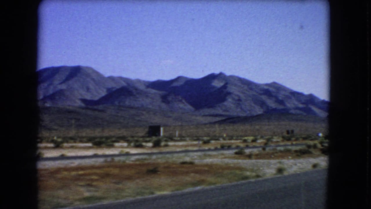 Las Vegas-area 1966 8mm frame showing roadside pavement, arid brush, a small dark structure, and distant mountain ridges.