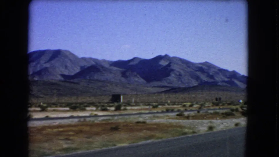 Las Vegas-area 1966 8mm frame showing roadside pavement, arid brush, a small dark structure, and distant mountain ridges.