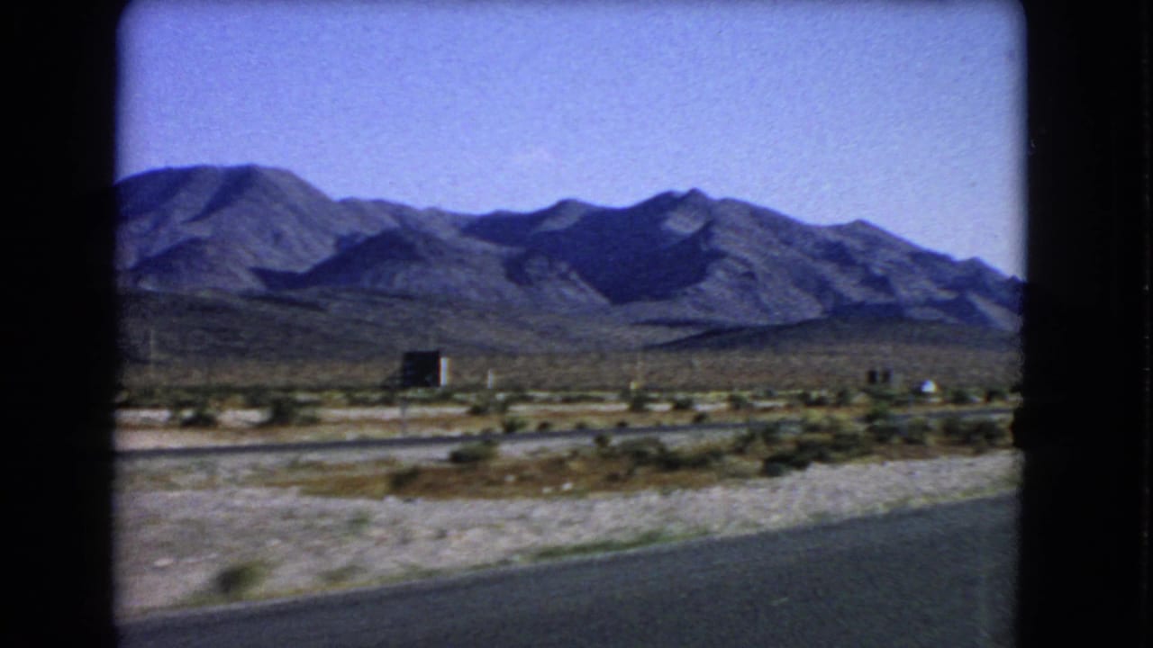 1966 Nevada travel frame with paved road, pale desert ground, sparse buildings or structures, and mountains beyond.