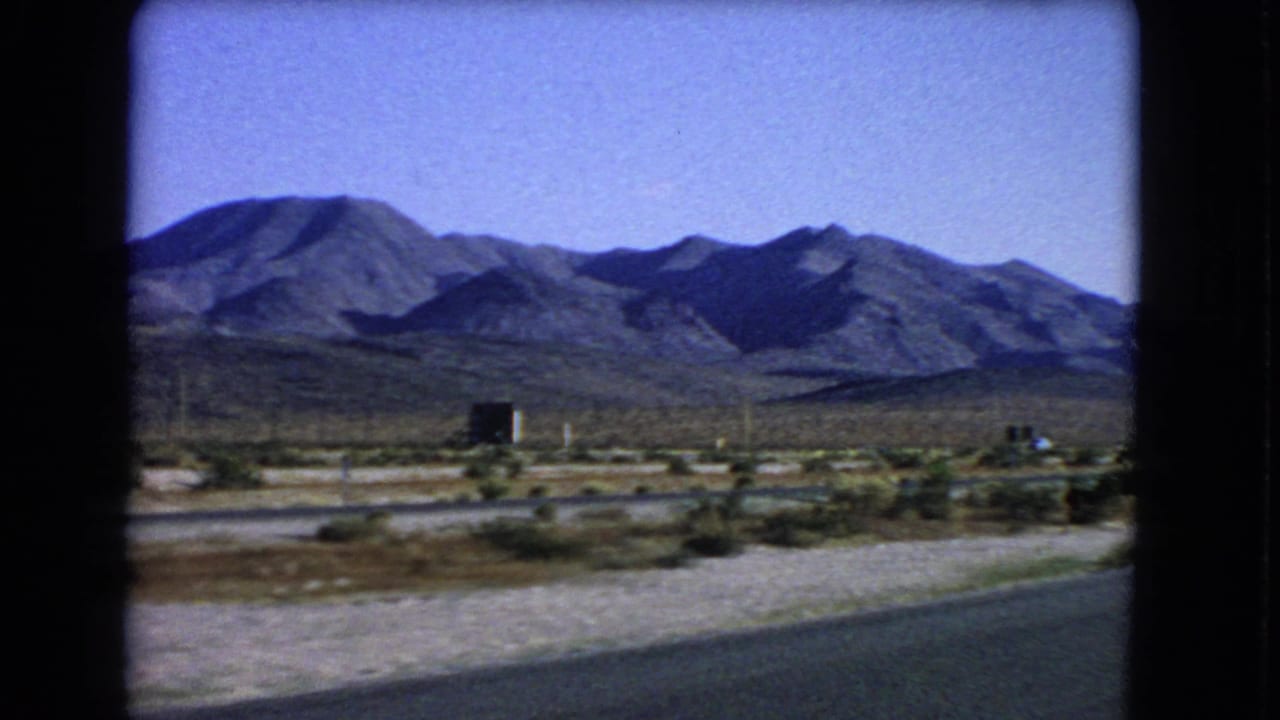 Desert roadside with rugged mountains under a blue sky, seen from a moving vehicle.