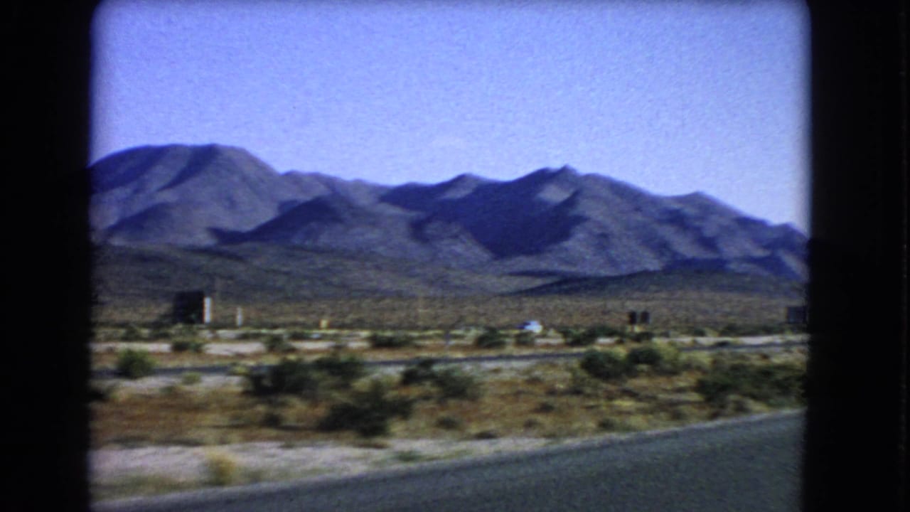 1966 Nevada driving view with desert brush, low roadside features, and shaded mountain slopes in the distance.