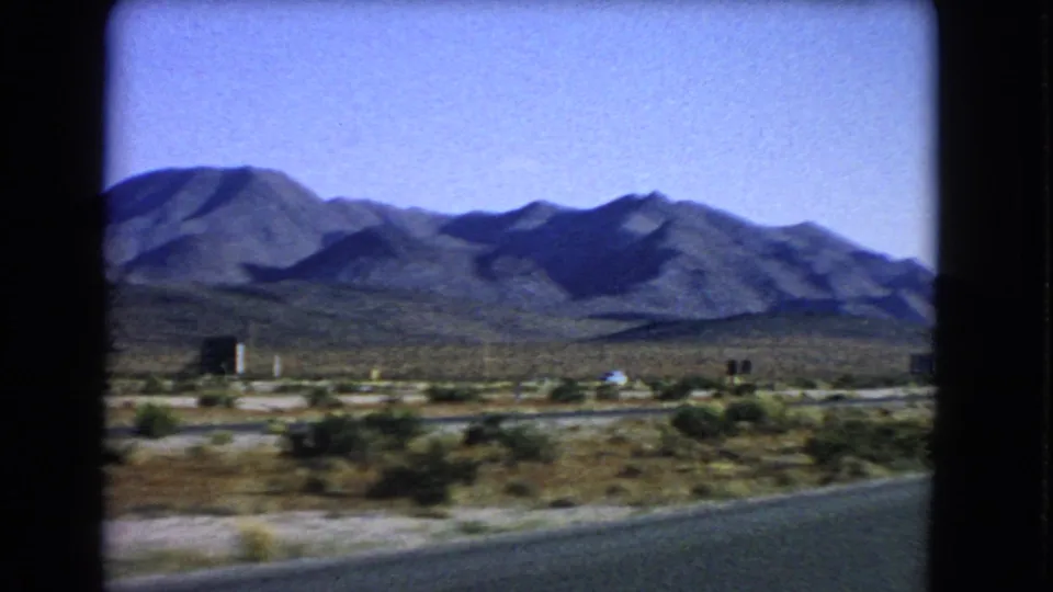 1966 Nevada driving view with desert brush, low roadside features, and shaded mountain slopes in the distance.