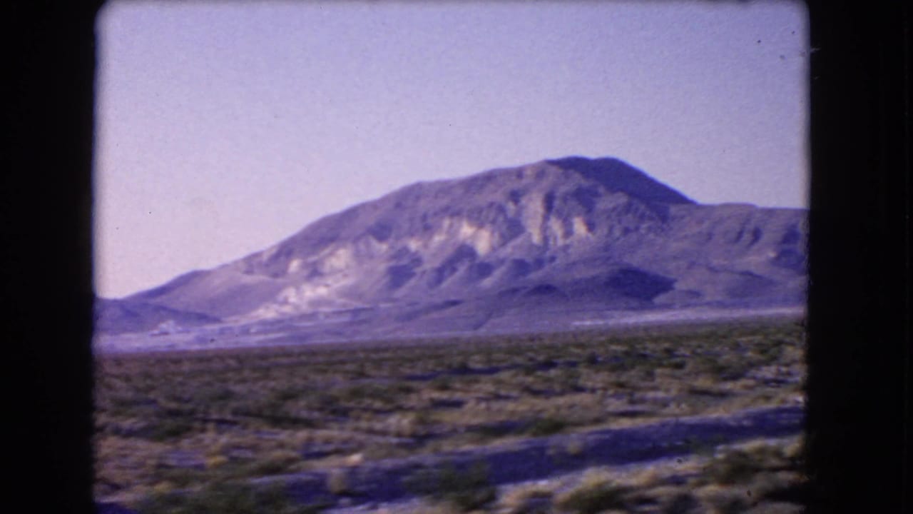 1966 Nevada desert frame showing a broad mountain mass, sparse scrubland, and a road edge from a moving vehicle.