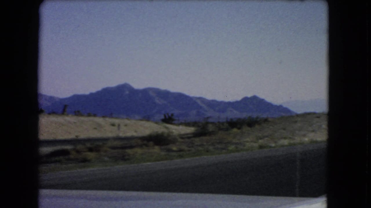 Moving-vehicle view near Las Vegas in 1966, with road pavement, low desert ground, and distant blue mountains under open sky.