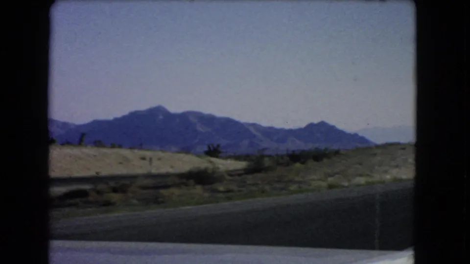 Moving-vehicle view near Las Vegas in 1966, with road pavement, low desert ground, and distant blue mountains under open sky.