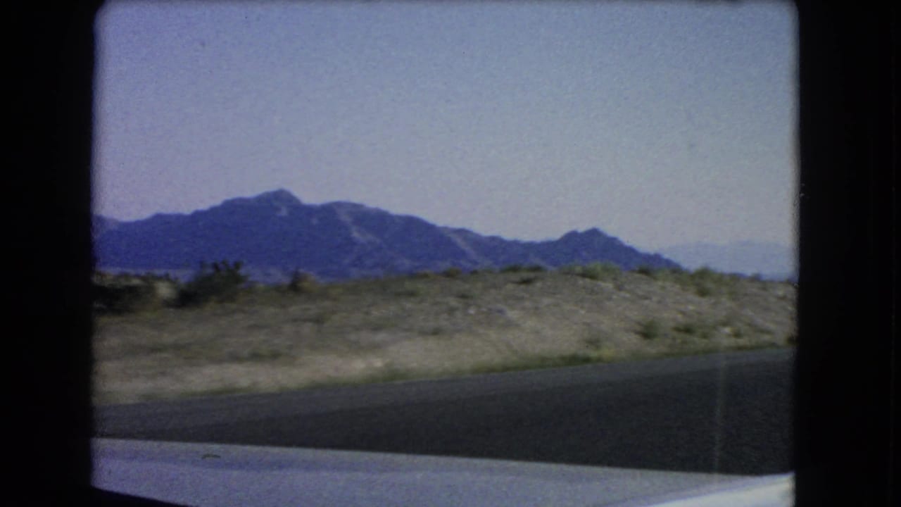 1966 Nevada 8mm frame showing roadside asphalt, scrubby desert foreground, and distant mountain peaks from a moving vehicle.