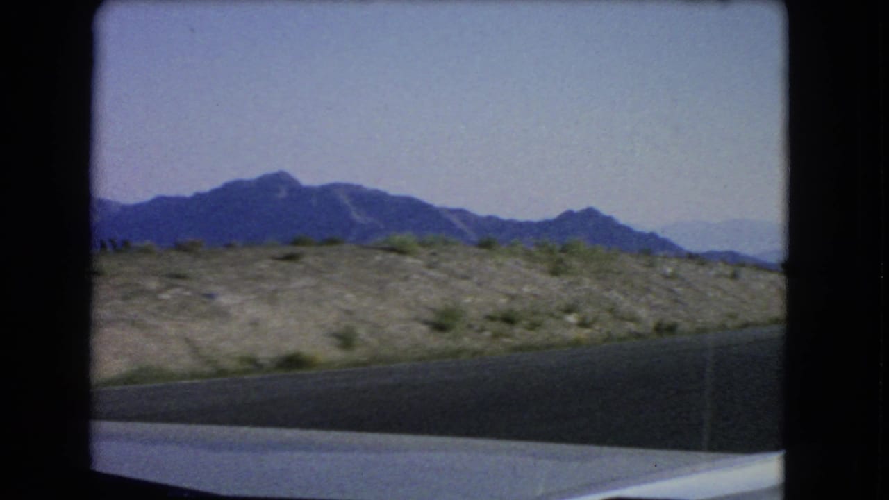 1966 8mm Las Vegas-area road view from a moving car, showing desert shoulder, scrub, distant mountains, and pale blue sky.