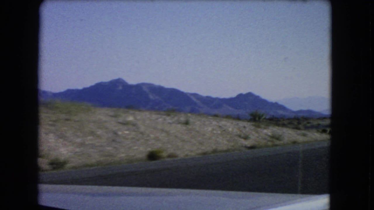 Desert roadway and rocky shoulder with blue mountains in the distance.
