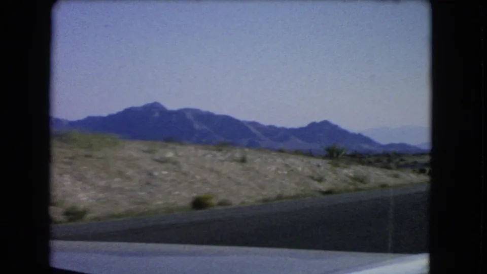 Passenger-side 8mm view near Las Vegas in 1966, with a dark road edge, scrubby desert ground, and mountain ridgeline.
