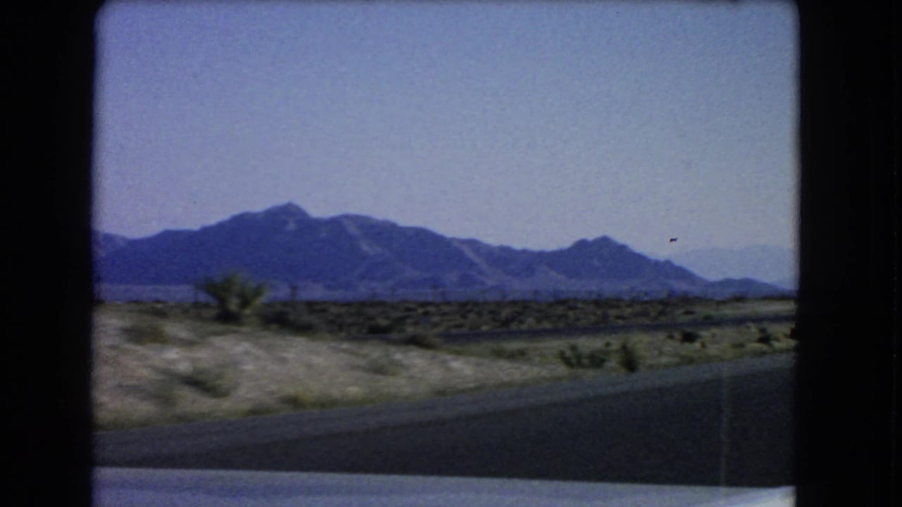1966 Nevada home-movie frame from a moving car, showing open desert, scattered shrubs, road foreground, and distant mountains.